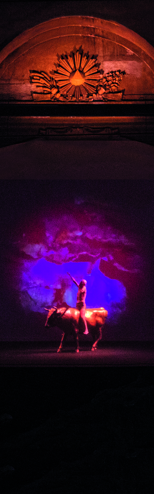 Un rosone dorato in alto con all'interno un simbolo sacro a forma di triangolo. Sotto una videoproiezione circolare inquadra una figura umana che cavalca a braccia aperte un toro dorato. Tutto il resto è nero.